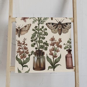 May include: A beige bath towel with a botanical and insect pattern. The design features brown moths, green plants in glass jars, and reddish-brown flowers. The towel is draped over a wooden ladder in a bathroom setting.
