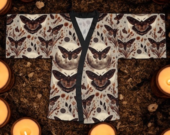 May include: A beige kimono with a repeating pattern of moths, moons, and botanical elements in shades of brown and tan. The kimono has a black trim and a V-neck design. The overall theme is a mystical or bohemian aesthetic.