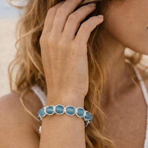 May include: A bracelet featuring a series of smooth, round, light blue sea glass beads connected by a white knotted cord. The beads are arranged in a single row, creating a continuous circular design that fits around the wrist securely.
