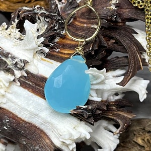 May include: A delicate gold chain necklace with a light blue teardrop-shaped gemstone pendant. The pendant is set in a simple gold wire wrap.