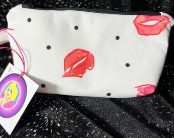 Può includere: Una pochette bianca con cerniera caratterizzata da un motivo a piccoli punti neri e labbra rosse. Un cordino rosa fissa un'etichetta quadrata bianca sul lato, che mostra un'illustrazione circolare di una persona con capelli biondi e il testo Rubella Vaxine.