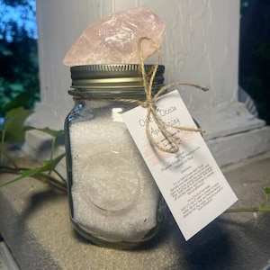 May include: A clear glass jar filled with white bath salts, topped with a large, light pink rose quartz crystal. The jar has a metal lid and a tag tied with twine that reads "One Ocean Apothecary".