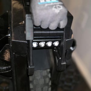 May include: Close-up of a black electric unicycle with a built-in LED light bar. The light bar has four bright white lights. The unicycle has a black tire and a black frame. A gloved hand is holding the handle.
