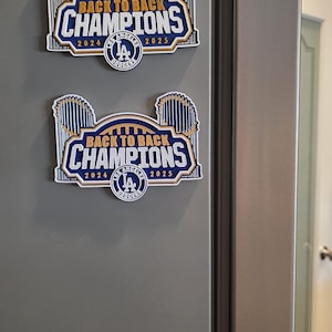 May include: Two magnets with a blue and white design, featuring the text "BACK TO BACK CHAMPIONS" and the years 2024 and 2025. The Los Angeles Dodgers logo is also visible. The magnets are attached to a gray surface.