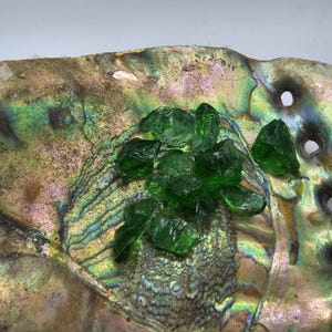 May include: Abalone shell with a shimmering, iridescent surface, displaying hues of green, gold, and purple. A cluster of faceted, emerald-green glass pieces rests on the shell, creating a contrast of textures and colors.