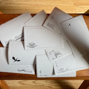 Könnte beinhalten: Eine Ansammlung weißer rechteckiger Karten mit schwarzen Strichzeichnungen von Büchern, Drachen und floralen Elementen. Die Karten liegen auf einer Holzoberfläche, passend für Schreibwaren oder Bastelarbeiten.