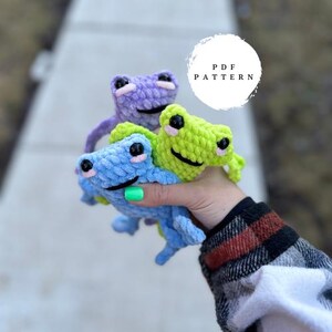 May include: Three crocheted frog plushies in different colours: purple, green, and blue. The frogs are holding onto each other and the text "PDF PATTERN" is visible in the background.