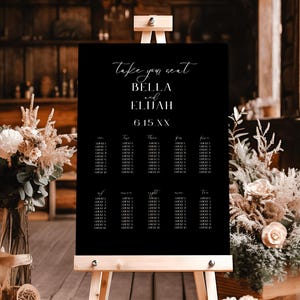 May include: Black wedding seating chart with white text. The chart is on an easel and says "Take your seat Bella and Elijah 6.15.XX".