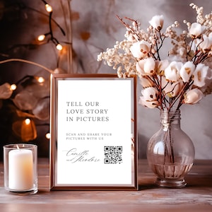 May include: A white sign with a gold frame on a wooden table. The sign says "Tell our love story in pictures. Scan and share your pictures with us. Camille and Theodore" and has a QR code.