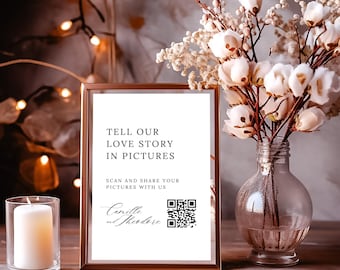 Può includere: Un cartello bianco con cornice dorata su un tavolo di legno. Il cartello dice "Tell our love story in pictures. Scan and share your pictures with us. Camille and Theodore" e ha un codice QR.