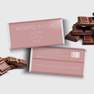 以下が含まれることがあります： ピンクのチョコレートバーの包装紙に「Wrapped in Love」という文字と「Victoria & Jackson」という名前が印刷されています。包装紙は白い縁取りと白いストライプで飾られています。包装紙の裏面にはバーコードと「Thank you for sharing our special day!」という文字が印刷されています。