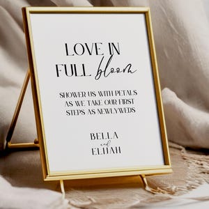 May include: A gold framed sign with the text "LOVE IN FULL bloom" and the names "BELLA and ELIJAH". The sign is on a stand, with a white background and black text. The sign is a wedding decoration.
