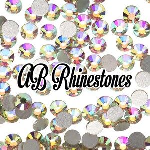 May include: A scattering of iridescent AB rhinestones and silver-backed rhinestones on a white background. The text "AB Rhinestones" is displayed in a black, cursive font.