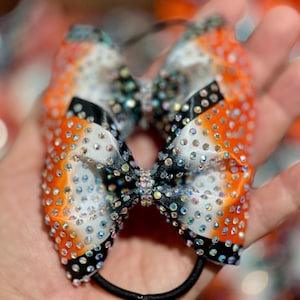 May include: A vibrant hair bow featuring a gradient of orange, white, and black, adorned with numerous iridescent rhinestones. The bow is attached to a black elastic band, creating a sparkling accessory.