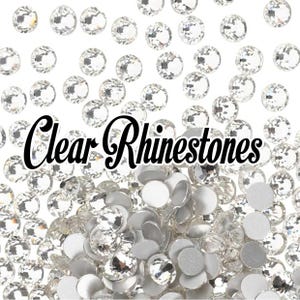 May include: A collection of clear rhinestones scattered on a white background. The image features a mix of faceted and flat-backed rhinestones, with the text "Clear Rhinestones" in a cursive font.