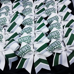 Könnte beinhalten: Eine Gruppe weißer und grüner Cheer-Schleifen mit dem aufgedruckten Text "HAWKS Cheer". Die Schleifen sind in einem Muster auf einer schwarzen Oberfläche angeordnet.