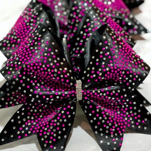 May include: Black cheerleading bows with a vibrant pink gradient and sparkling rhinestones. The bows have a satin finish and a rhinestone-studded center. These accessories are perfect for cheerleaders and dancers.