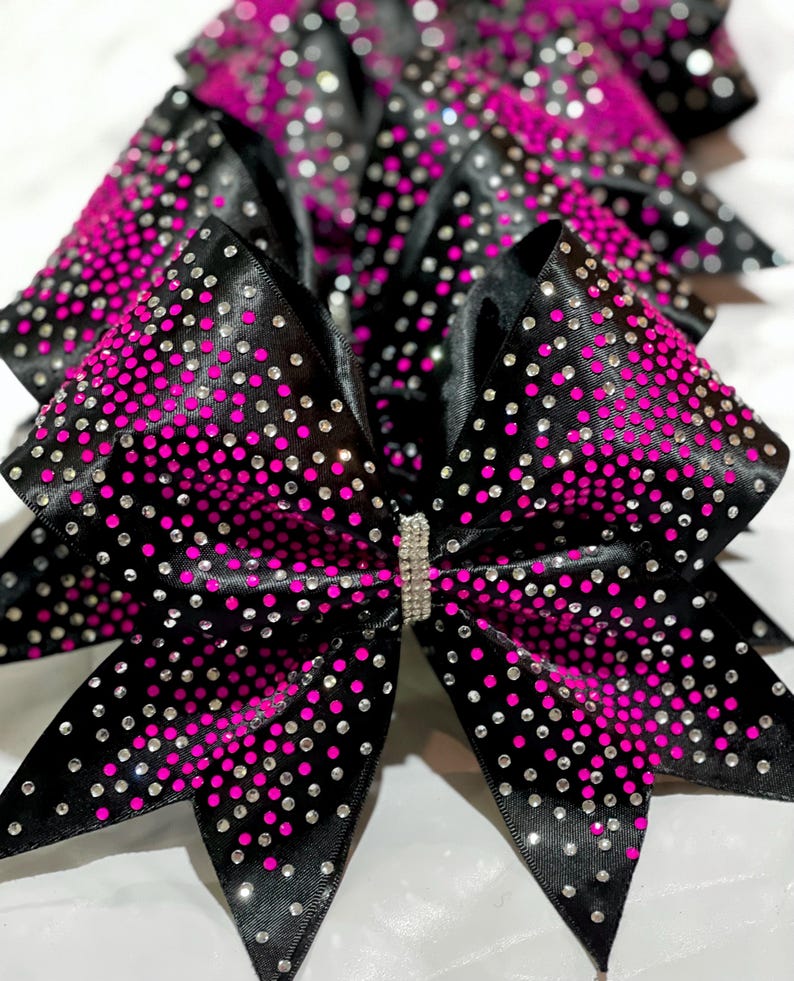 May include: Black cheerleading bows with a vibrant pink gradient and sparkling rhinestones. The bows have a satin finish and a rhinestone-studded center. These accessories are perfect for cheerleaders and dancers.