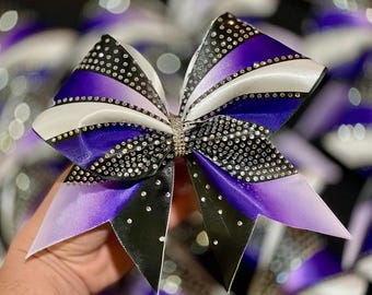Puede incluir: Un gran lazo de animadora morado, blanco y negro con pedrería. El lazo está hecho de cinta de satén y tiene un efecto degradado.