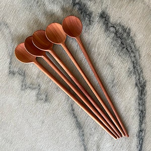 May include: Set of five long wooden spoons with a reddish-brown color. The spoons are arranged in a fan shape, with the handles aligned at the bottom.