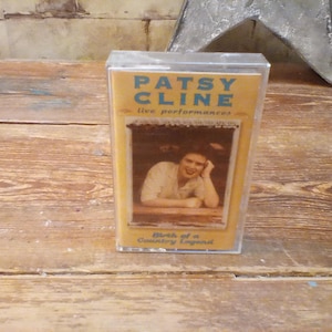 Op de afbeelding: Een vintage cassettebandje met Patsy Cline's "Live Performances". De gele en blauwe hoes toont de naam van de artiest en een foto. De tekst "Birth of a Country Legend" is ook zichtbaar.