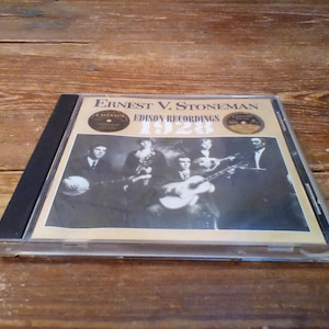 Peut inclure: Un boîtier de CD avec l'album "Ernest V. Stoneman 1928 Edison Recordings". La couverture présente une photographie en noir et blanc d'un groupe jouant des instruments. Le boîtier est transparent avec un dos noir, posé sur une surface en bois.