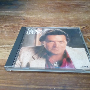 以下が含まれることがあります： 「The Very Best of Mickey Gilley」のアルバムが収録されたCDケース。ジャケットには、スーツジャケットと赤と白のチェック柄のシャツを着た男性の肖像画が描かれています。CDケースは黒と透明で、木製の表面に置かれています。
