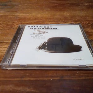 Può includere: Custodia per CD con l'album "Sonny Boy Williamson - The Real Folk Blues". La copertina presenta un cappello nero e un'armonica su sfondo bianco. La custodia del CD è trasparente e il titolo dell'album è stampato in nero.