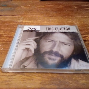 Peut inclure: Un boîtier de CD présentant l'album d'Eric Clapton, "The Millennium Collection". La couverture affiche un portrait en noir et blanc de Clapton. Le boîtier du CD est transparent, et le titre de l'album est imprimé en blanc.