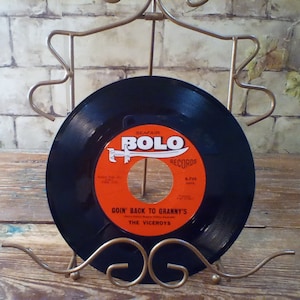 Puede incluir: Un disco de vinilo negro con una etiqueta roja. La etiqueta dice "Seafair Bolo Records" y "Goin' Back To Granny's" por "The Viceroys". El disco está en un soporte de metal dorado.