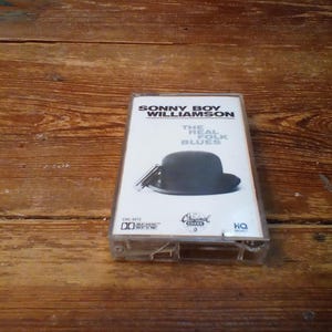 Può includere: Una musicassetta vintage dell'album "The Real Folk Blues" di Sonny Boy Williamson. La copertina presenta un cappello a bombetta nero e un'armonica. La cassetta è in un astuccio di plastica trasparente, su una superficie di legno.