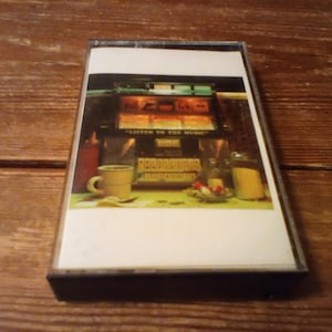 May include: A rectangular cassette tape case featuring a white cover with a central photograph of a vintage jukebox, a light-colored mug, and a glass container. The text "LISTEN TO THE MUSIC" is printed in white on the front of the jukebox image.