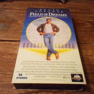 May include: A VHS tape of the film Field of Dreams featuring Kevin Costner on the cover. The artwork shows a person standing in a field with a blue sky background. The text includes the title, cast, and production credits in black font.