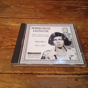 May include: A CD case featuring the album "Memphis Minnie & Kansas Joe, Volume 4, 1933-1934." The cover is black and white, with portraits of the artists and text. The CD case is on a wooden surface.