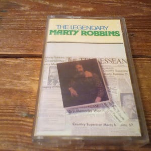 May include: A clear plastic cassette tape case featuring a white insert with the text The Legendary Marty Robbins in green and blue. The insert displays a newspaper collage titled The Tennessean with a central photograph of a man in a patterned shirt.