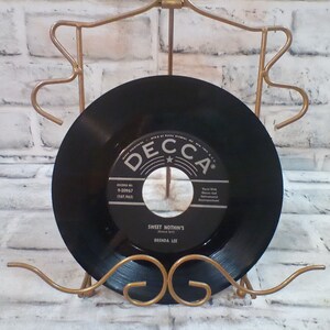 Peut inclure: Un disque vinyle noir avec l'étiquette "Decca" et le titre "Sweet Nothin's" de Brenda Lee. Le disque est sur un support en métal doré.