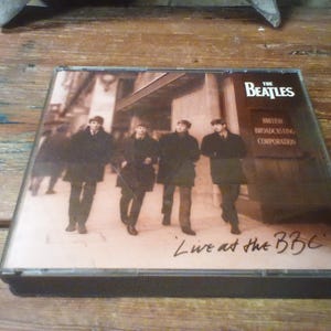 May include: A square CD case featuring The Beatles Live at the BBC album cover. The artwork displays a sepia-toned photograph of the four band members walking outdoors near a building labeled British Broadcasting Corporation in white text.