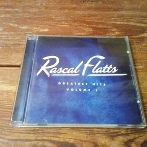 May include: A Rascal Flatts Greatest Hits Volume 1 music CD in a clear plastic jewel case. The cover art features a deep blue, textured background with the band name in white script and the album title in white sans-serif capital letters.