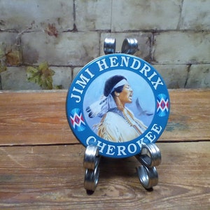 Peut inclure: Un bouton rond bleu et blanc avec une image de Jimi Hendrix portant une coiffe amérindienne. Le bouton porte l'inscription "Jimi Hendrix Cherokee" sur le pourtour.
