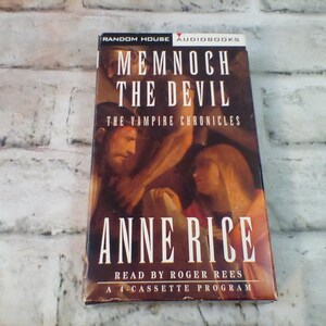 Puede incluir: Una caja de cassette roja y blanca con el t&iacute;tulo "Memnoch the Devil" de Anne Rice. La caja presenta una pintura de un hombre y una mujer, y el texto "The Vampire Chronicles".