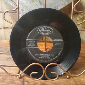Könnte beinhalten: Eine schwarze 45-U/min-Schallplatte der Platters mit dem Titel Smoke Gets In Your Eyes auf dem Label Mercury Records. Die runde Platte hat ein schwarzes Mittellabel mit weißer Schrift und ein großes Mittelloch, präsentiert auf einem kupferfarbenen Metallständer.