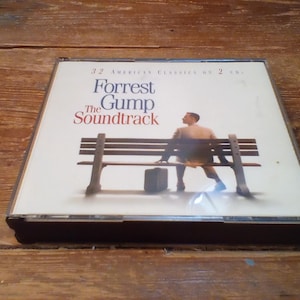 May include: A CD case for "Forrest Gump: The Soundtrack" featuring a man sitting on a park bench. The cover art includes the film's title and the text "32 American Classics on 2 CDs."