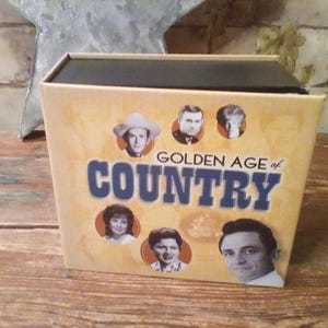 Puede incluir: Una caja cuadrada titulada Golden Age of Country con fondo color canela. El frente presenta texto azul y seis retratos circulares en blanco y negro de varios artistas de música country, incluido un retrato destacado de Johnny Cash abajo.