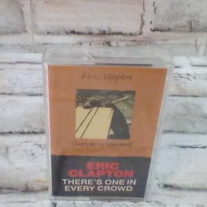 May include: A cassette tape with a brown background and a black and white photo of a dog lying on a bench. The text "Eric Clapton There's One in Every Crowd" is printed in red and white on the front of the cassette tape.