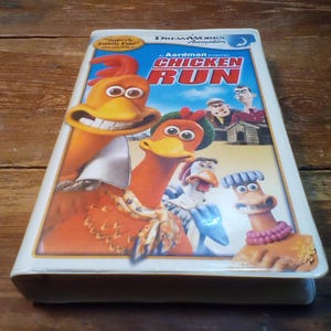 Puede incluir: Una cinta de vídeo VHS de la película de animación "Chicken Run" con pollos de dibujos animados en la portada. El título está en letras rojas grandes, con los logotipos de DreamWorks y Aardman Animation visibles. La cinta es blanca con una imagen colorida.