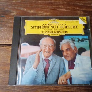 May include: A Deutsche Grammophon CD case featuring Aaron Copland's Symphony No. 3 and Quiet City, performed by the New York Philharmonic under Leonard Bernstein. The cover shows a photograph of two men, one wearing a striped blazer and red tie.