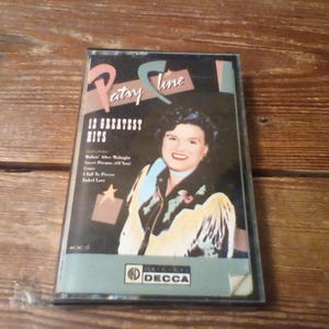 May include: A Patsy Cline 12 Greatest Hits cassette tape in a clear plastic case. The cover features a portrait of the artist wearing a fringed jacket, with the text Patsy Cline, 12 Greatest Hits, and a list of songs including Walkin' After Midnight.