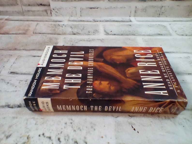 Puede incluir: Un libro de bolsillo titulado "Memnoch el Diablo" de Anne Rice. El libro forma parte de la serie "Las Cr&oacute;nicas Vamp&iacute;ricas" y est&aacute; publicado por Random House Audiobooks. La portada presenta una pintura de dos personas abraz&aacute;ndose.