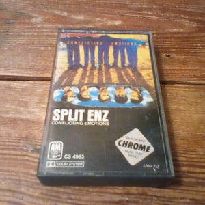 May include: A cassette tape of the album Conflicting Emotions by Split Enz. The cover features a group of people standing on a sandy surface with their reflections visible in a pool of water below. The text reads Split Enz, Conflicting Emotions, A&M, CS 4963, and Chrome Music That Shines.