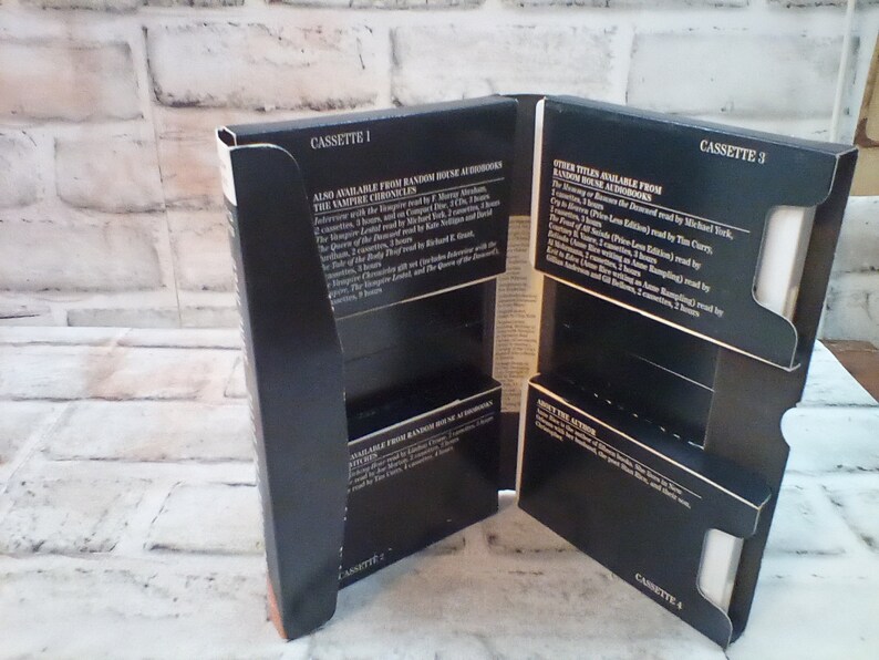 Puede incluir: Cuatro cajas de cassette negras con texto blanco. El texto en las cajas dice "Cassette 1", "Cassette 2", "Cassette 3" y "Cassette 4". Las cajas est&aacute;n apiladas una encima de la otra. El texto en las cajas tambi&eacute;n enumera los t&iacute;tulos disponibles de Random House Audiobooks, incluyendo "The Vampire Chronicles" y "The Queen of the Damned".